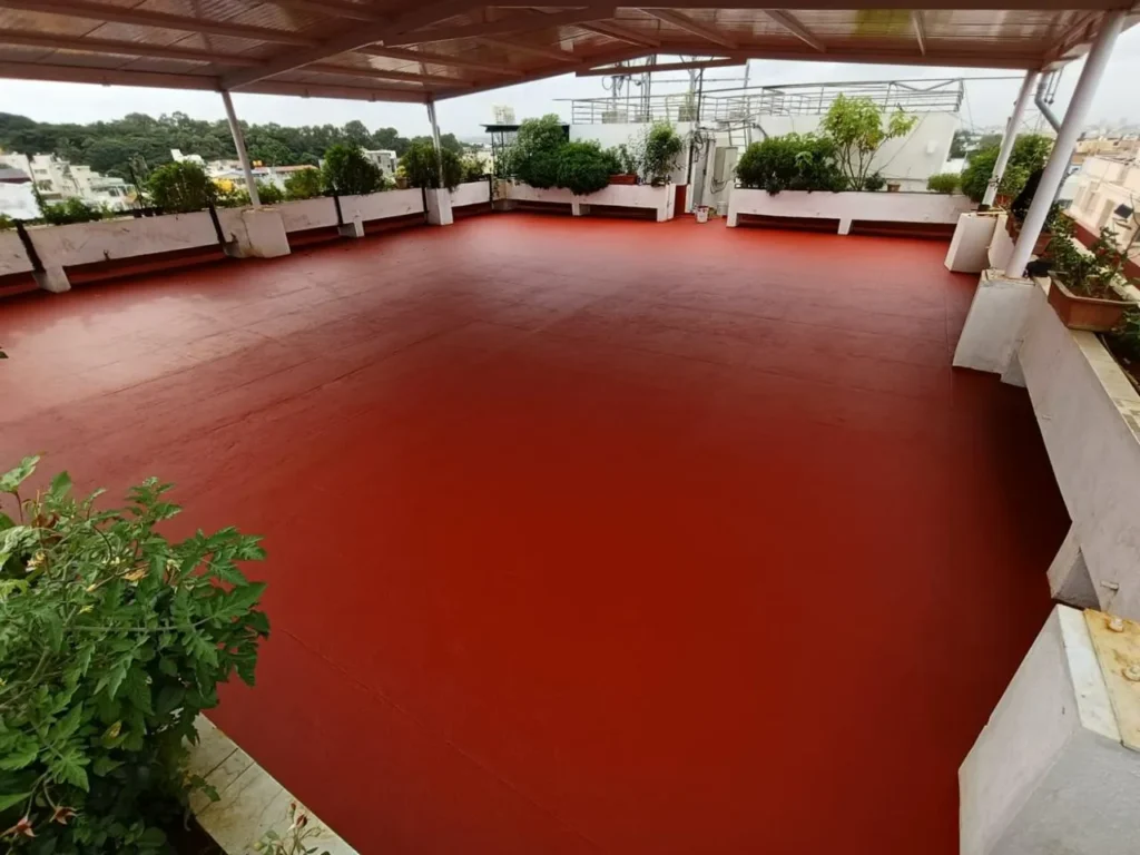 A rooftop patio with a red floor and a white overhead canopy, surrounded by white planters and green trees, under an overcast sky. Paint buckets and a roller are visible on the floor.